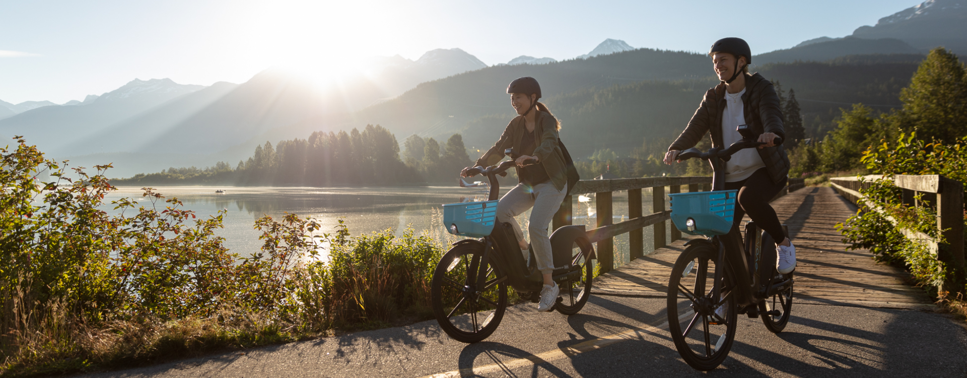 Two people, a young man and young woman, ride BCAA's Evolve e-bikes on a paved trail in Whistler.