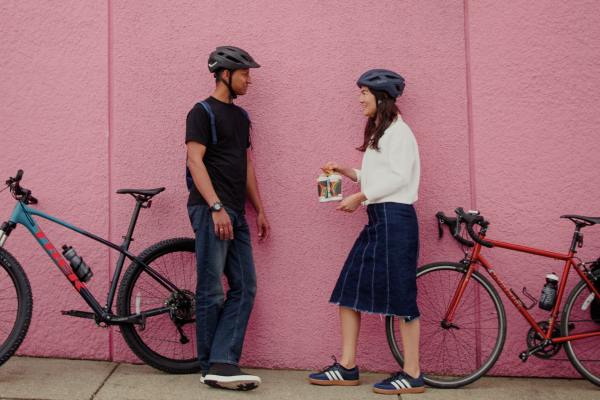 A man and a woman stand with their bikes in front of a pink wall. Both are wearing helmets. The woman holds a case of beer.