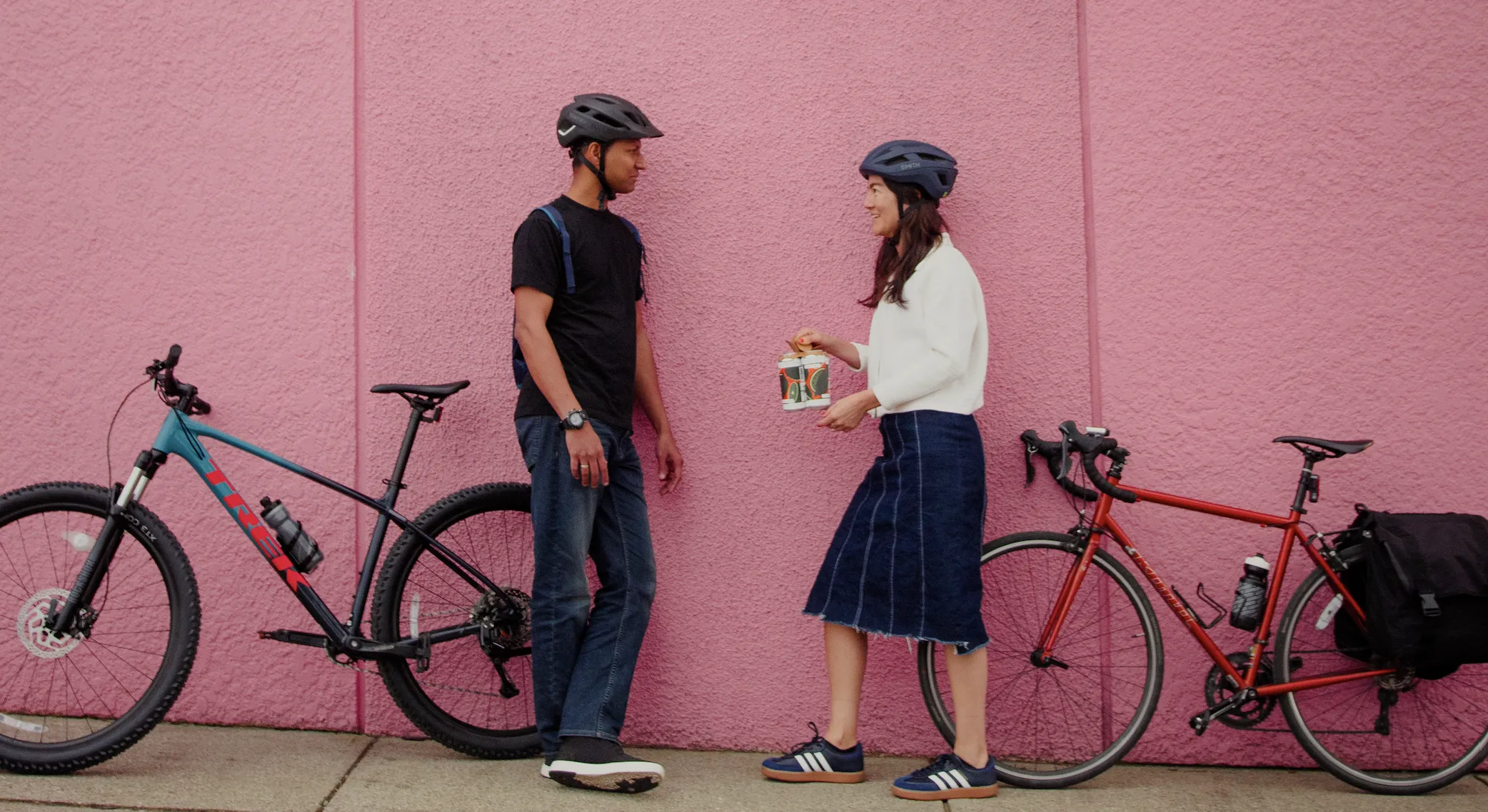 Two people stand against a pink wall with their bikes behind them. One woman is holding a 4-pack of beer.